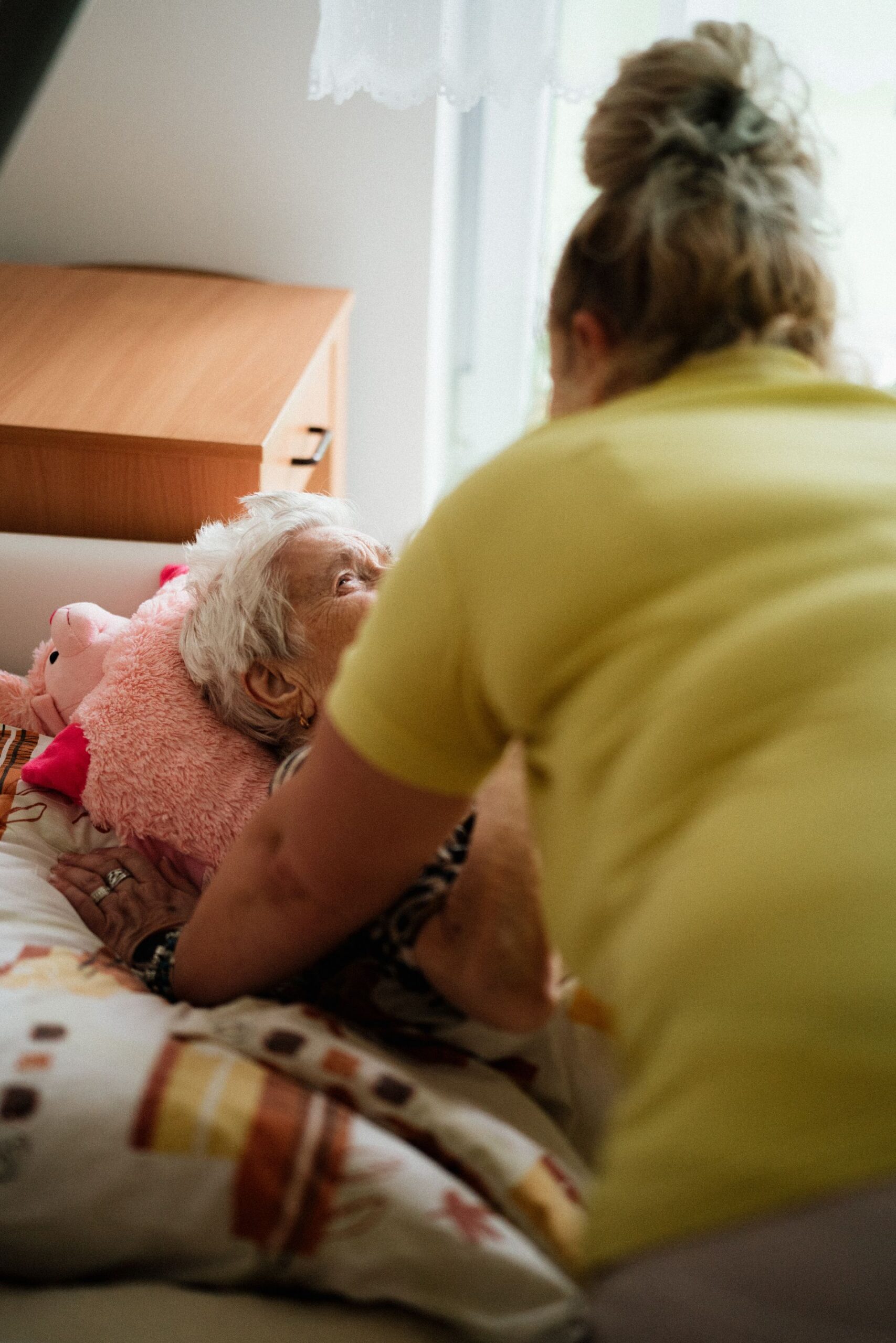 We zien een mevrouw van de achterkant. De mevrouw leunt over een bed. In het bed ligt een mevrouw op leeftijd. Ze kijken elkaar aan en de mevrouw op leeftijd lacht.